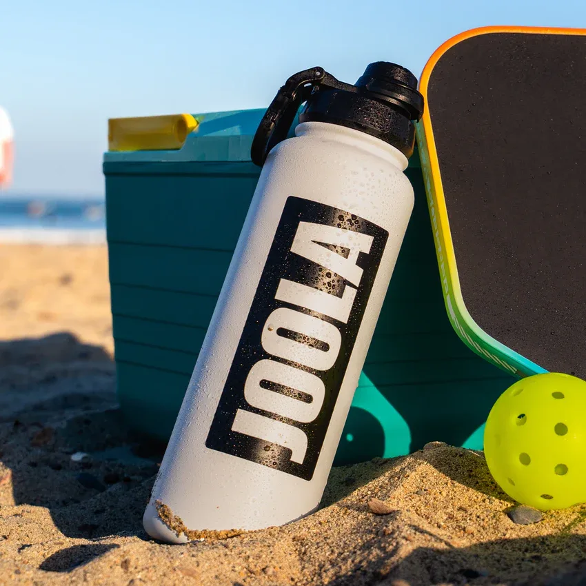 Bình nước JOOLA 40 oz dựng nghiêng cạnh thùng giữ lạnh và vợt pickleball trên bãi cát